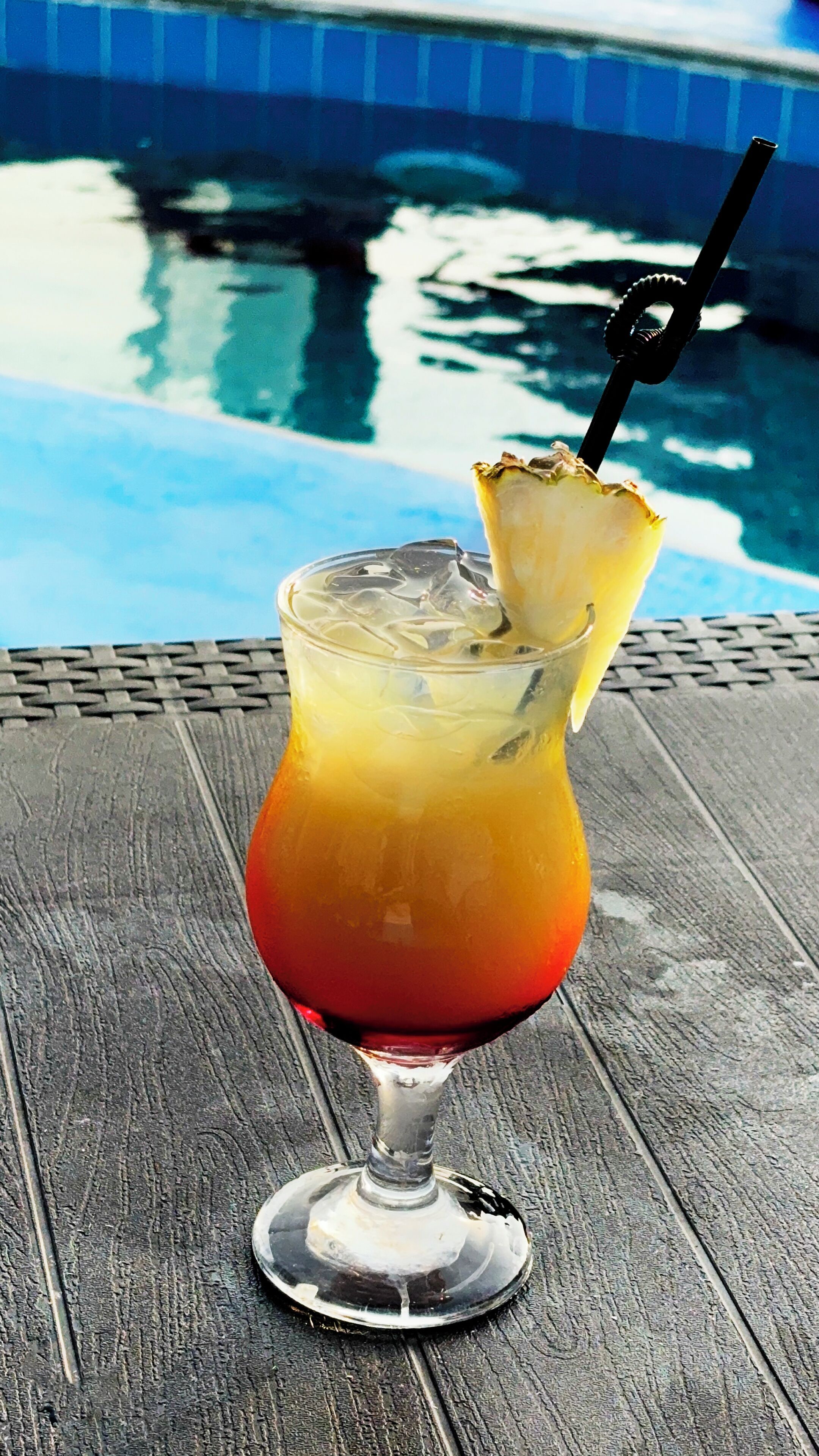 A cocktail glass with a slice of pineapple on a wooden table by a pool.