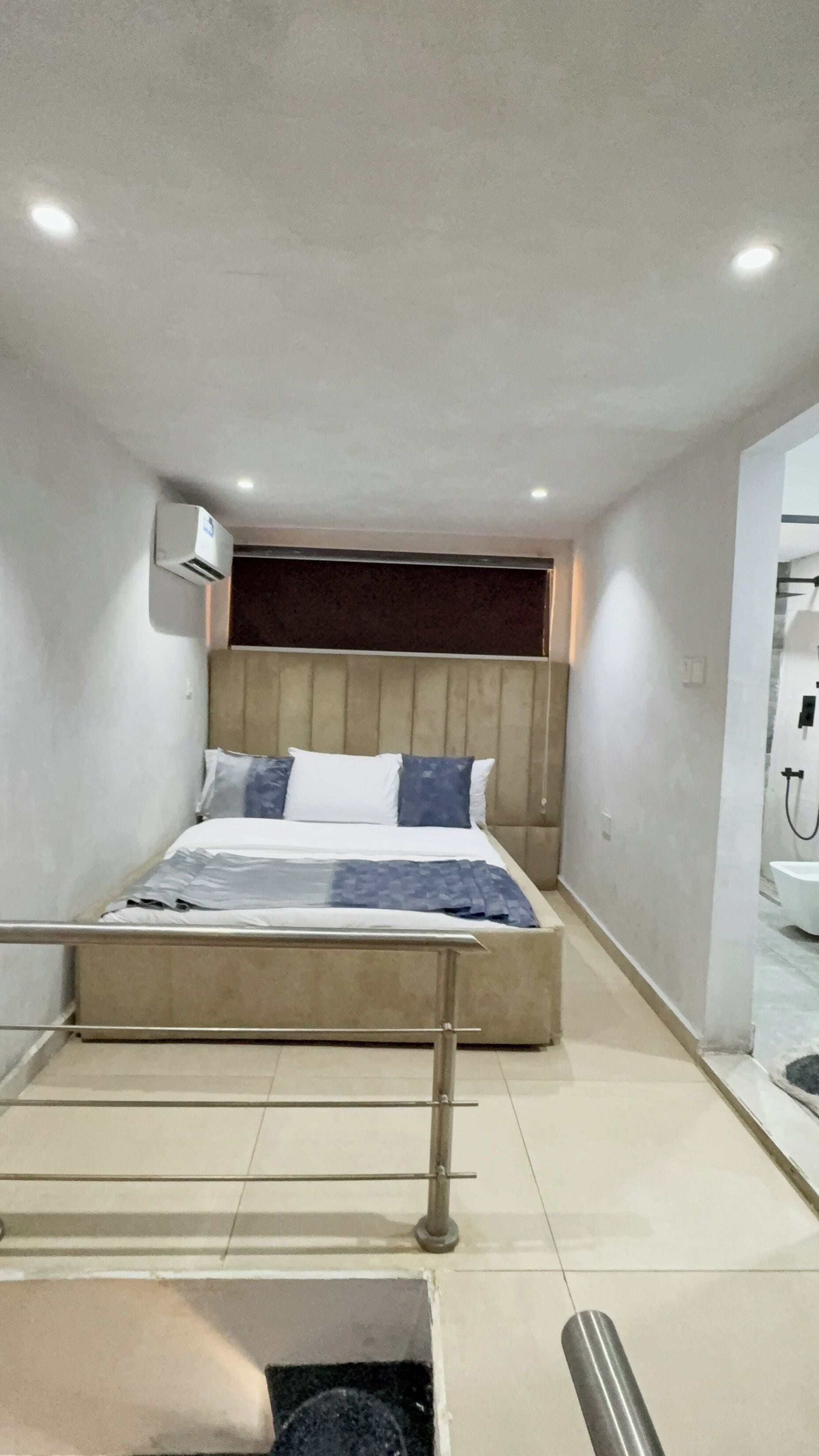 Una habitación de hotel moderna con una cama, un cabecero, un aire acondicionado de pared y un baño con lavamanos y espejo.