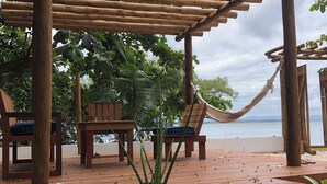 Eine Holzterrasse mit Stühlen und einer Hängematte, umgeben von viel Grün und Blick aufs Meer.