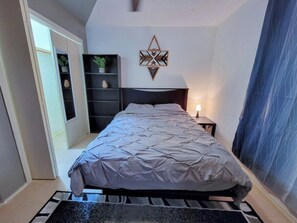 A bedroom with a bed, a nightstand, a wall-mounted decorative piece, a shelving unit with plants, and a window. - Entire house/Large TVs in all rooms/ Massage Chair/Gym/Grill (Houston)