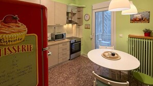 A compact kitchen with a red refrigerator, a microwave, and a stove. There is a small dining table with chairs. The walls are painted green, and there is a window with a view of a balcony. - Miró by halu! - Cozy apartment on Mitropoleos street (Thessaloniki)