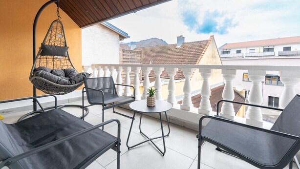 A balcony with a hanging chair, two black chairs, a small round table with a plant, and a view of buildings and mountains. - Large Maisonette 90 m² | For Groups | 9 Guests (Kapfenberg)
