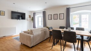 A modern living room with a sofa, dining table, and chairs. There is a flat-screen TV mounted on the wall, a radiator, and a window with curtains. - Large Maisonette 90 m² | For Groups | 9 Guests (Kapfenberg)