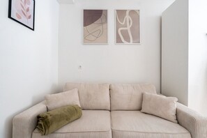 Un sofá beige con textura y una manta verde, dos cuadros abstractos en la pared, y una pared blanca.