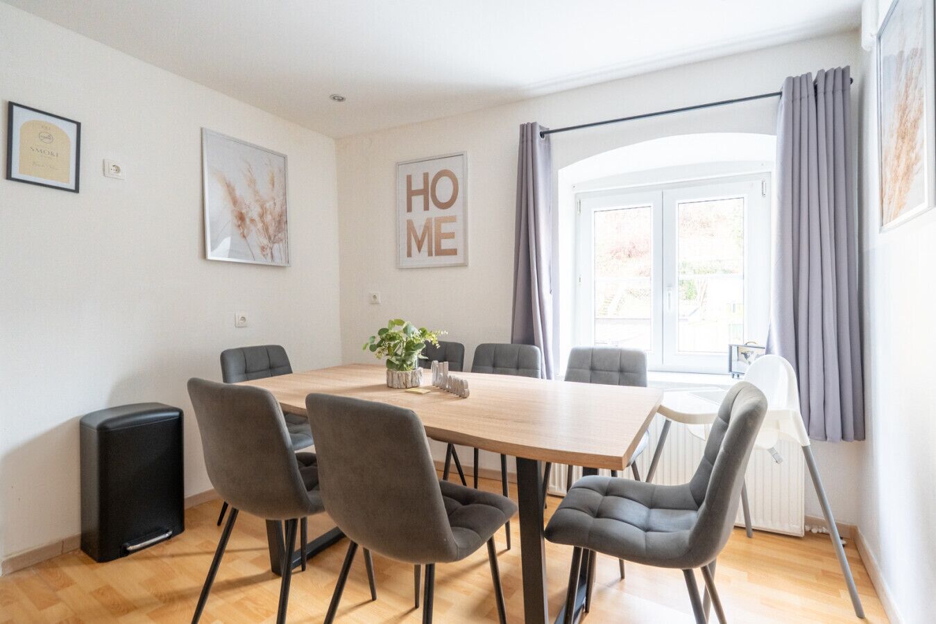 Un comedor moderno con una mesa de madera y sillas tapizadas en gris, un pequeño centro de mesa con planta y un amplio ventanal con cortinas.