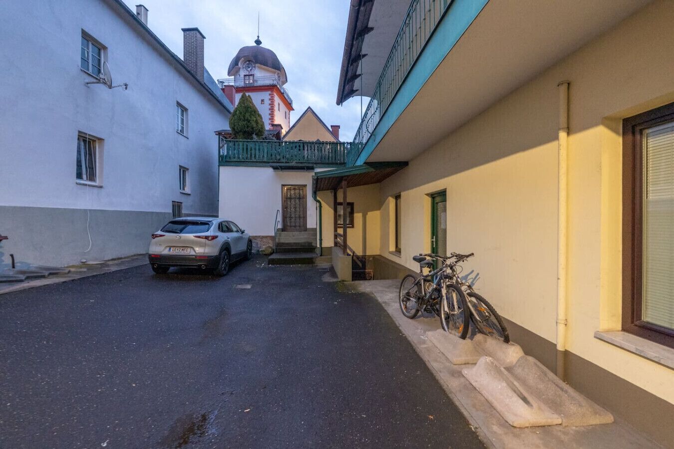 Eine schmale Straße mit einem abgestellten Auto und einem Fahrrad, das an einem Gebäude lehnt.