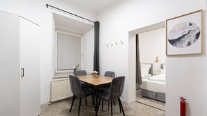 Ein Hotelzimmer mit einem Holztisch, vier Stühlen, einem Fenster mit Jalousien, einem Bett mit weißer Bettwäsche, einem gerahmten Bild an der Wand und einem Feuerlöscher.