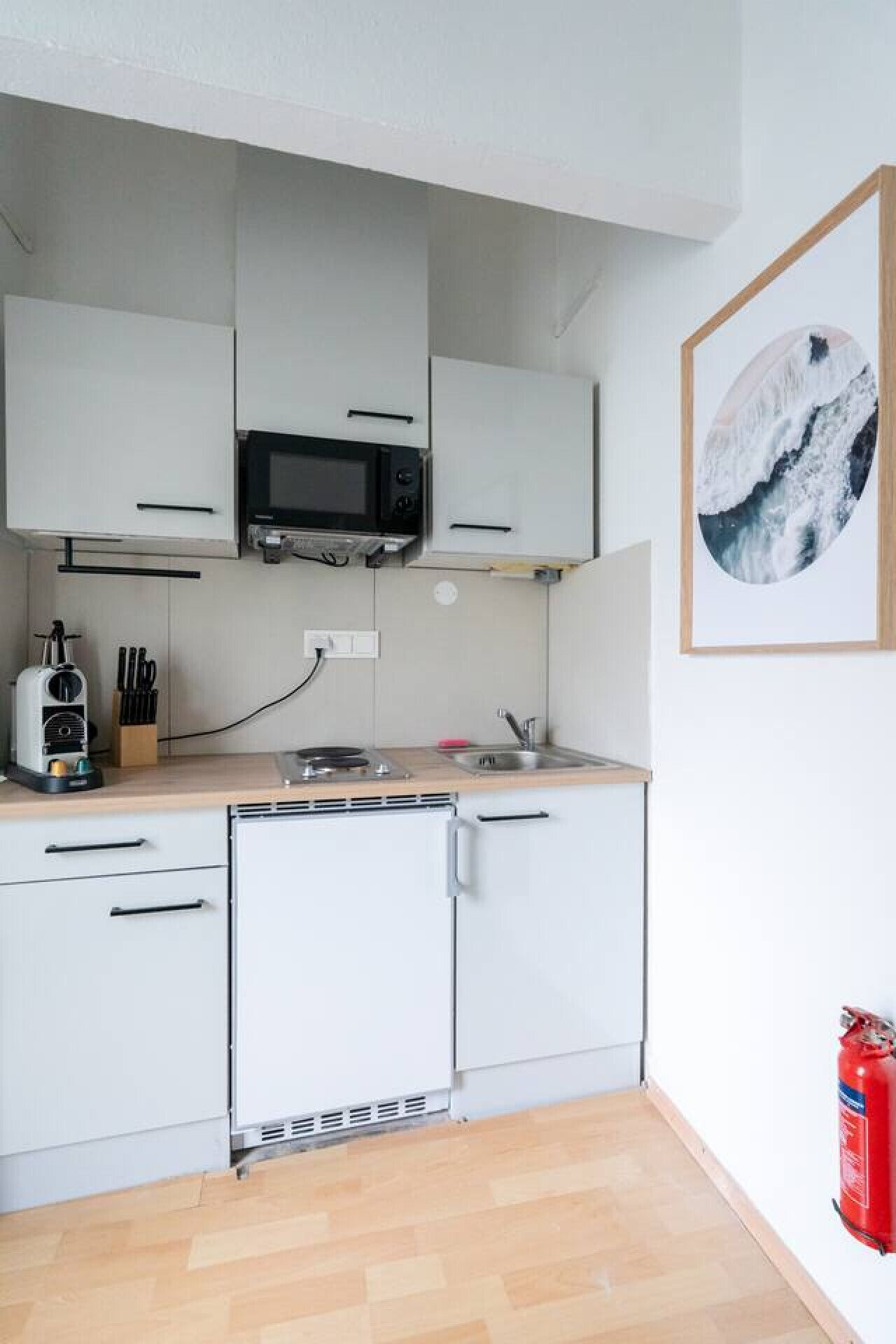 A compact kitchen with white cabinets, a microwave, a toaster, and a sink. There is a fire extinguisher on the floor.
