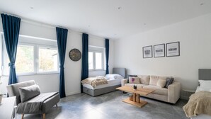 A modern living room with a sofa, a bed, a coffee table, and a couch. There are three windows with blue curtains, a round wall clock, and motivational posters on the wall. - 60m² Apt • Business & Family Ready • Near Vienna (Purkersdorf)