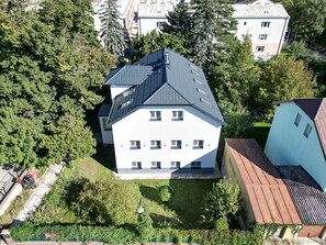 Um edifício de dois andares com telhado inclinado, cercado por árvores e edifícios vizinhos.