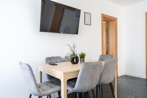 Ein moderner Essbereich mit einem Holztisch, vier grau gepolsterten Stühlen, einem Blumentopf und einem angebrachten Flachbildfernseher.