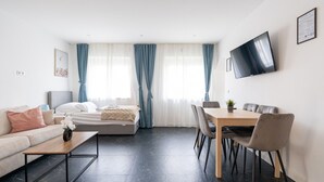 Una habitación de hotel moderna con cama, sofá, mesa de comedor con sillas, televisor montado en la pared y ventana con cortinas.