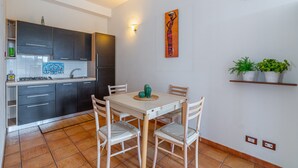 A compact kitchen with dark cabinets, a white dining table, and chairs, a wall-mounted light, and a potted plant on a shelf. - Apartment 'Pathos Retreat Mandorlo' with Shared Pool, Wi-Fi and Air Conditioning (Arenella)