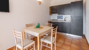 A compact kitchen with dark cabinets, a tiled backsplash, a dining table with chairs, and a wall-mounted light fixture. - Apartment 'Pathos Retreat Ulivo' with Shared Pool, Wi-Fi and Air Conditioning (Arenella)
