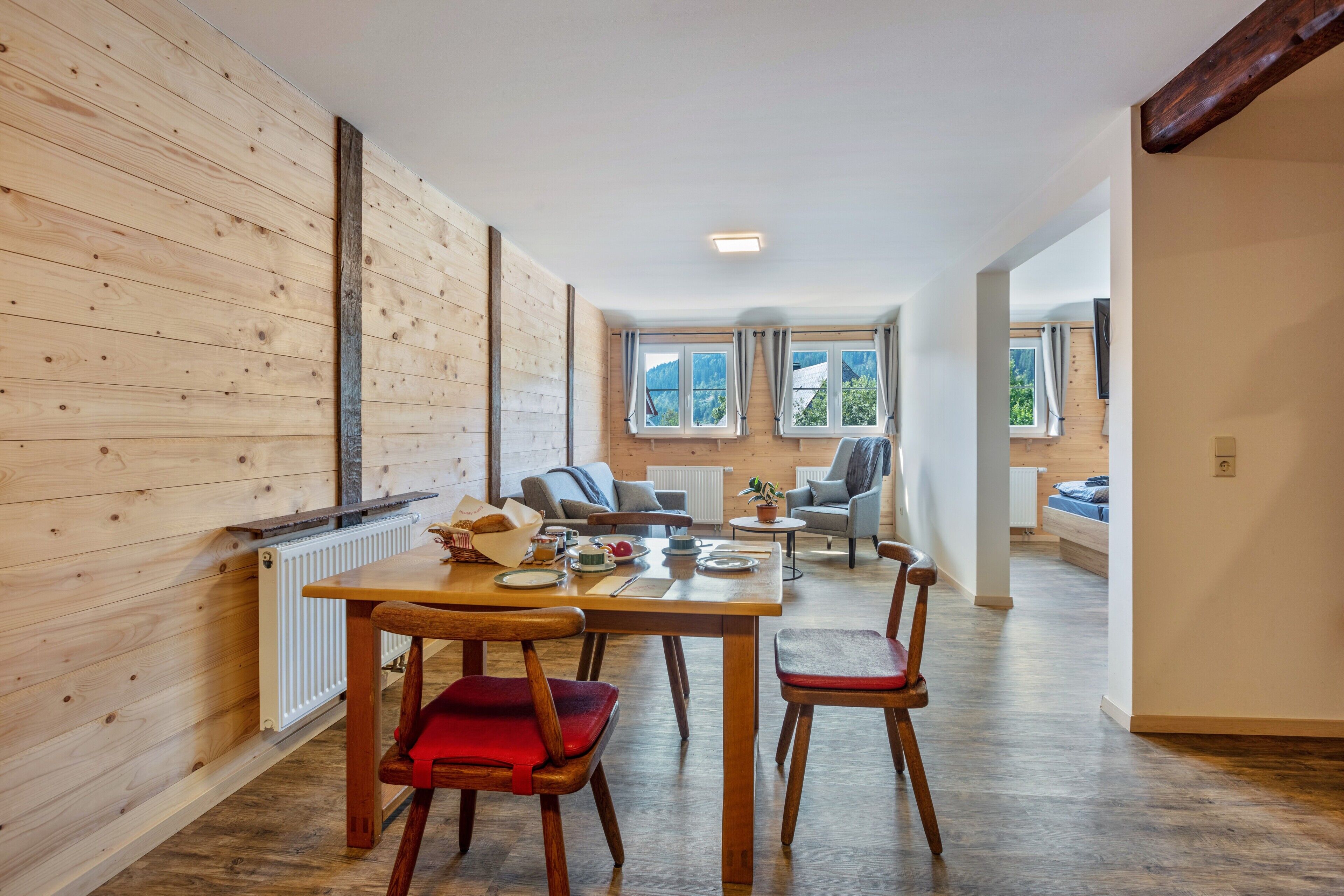 Une pièce aux murs lambrés, avec une table à manger dressée pour quatre personnes, un radiateur et une vue sur l’extérieur grâce à de grandes fenêtres.
