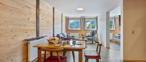 Een kamer met houten lambrisering, een gedekte eettafel voor vier personen, een radiator en uitzicht op het buitenlands landschap door grote ramen.