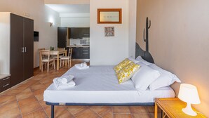A bedroom with a bed, a bedside table with a lamp, a wardrobe, a dining area with chairs, and a kitchen in the background. - Apartment 'Pathos Retreat Zagara' with Shared Pool, Wi-Fi and Air Conditioning (Arenella)