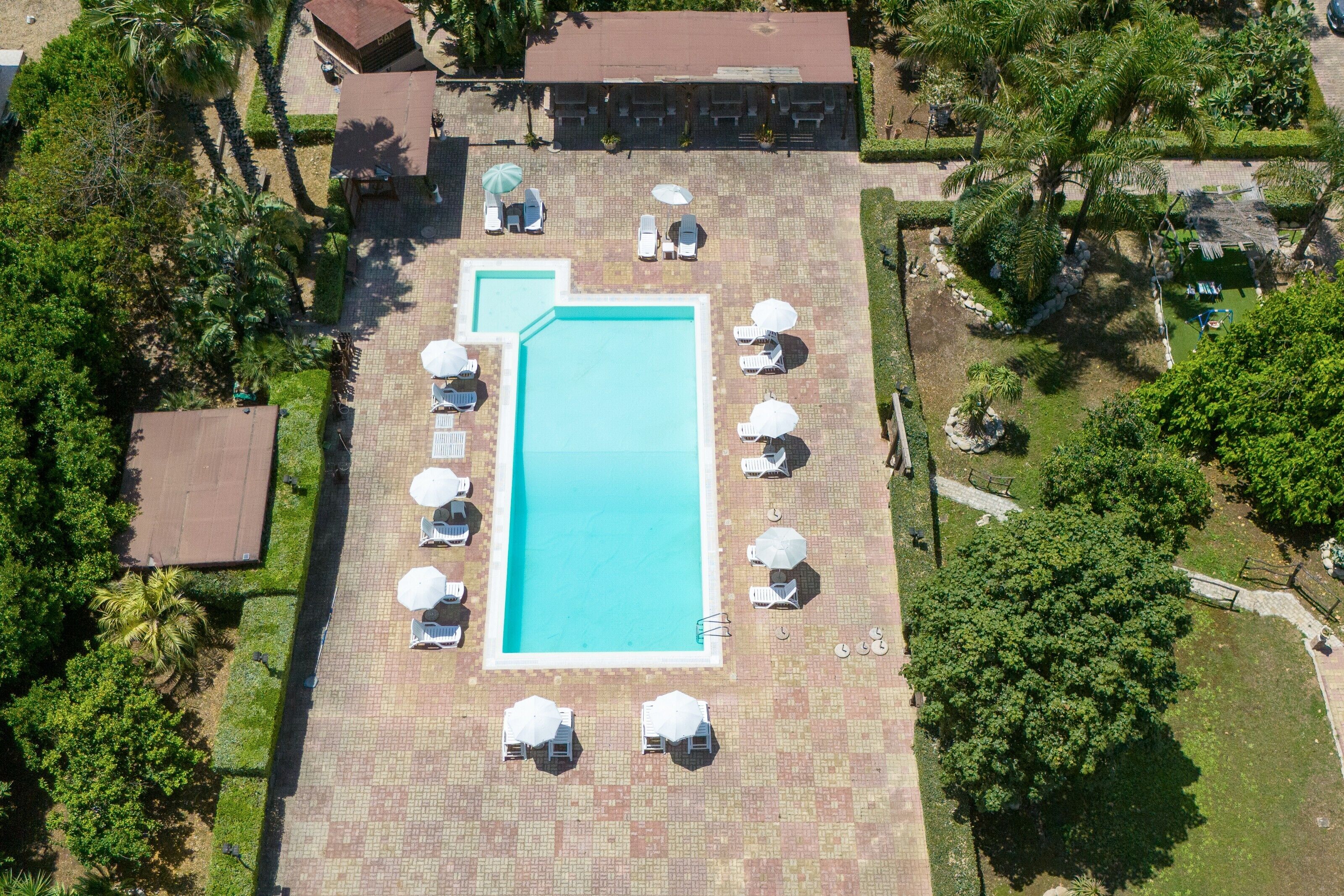 Ett flygbildsfoto av ett semesterområde med en rektangulär pool omgiven av solstolar och parasoller, med palmer och en byggnad i bakgrunden.