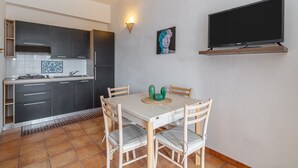Uma cozinha compacta com armários escuros, uma mesa de jantar branca com quatro cadeiras, uma televisão de tela plana na parede e um pequeno cacto decorativo na mesa.