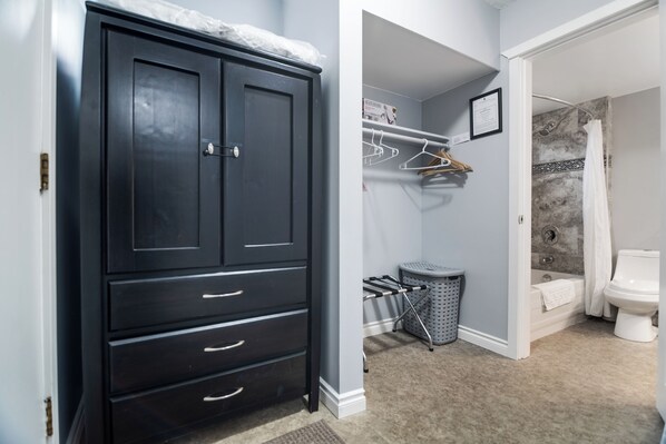 A room with a dark wooden wardrobe, a walk-in closet with hanging clothes, a grey trash bin, and a bathroom with a bathtub and toilet. - #201 Lower Garden View Suite at Shorewater Resort, Qualicum Beach (Qualicum Beach)