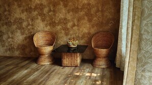 A room with a rustic wooden floor, two wicker chairs, a small table with a basket, and a patterned curtain. - Beltola Lodge - a serene homestay (Guwahati)