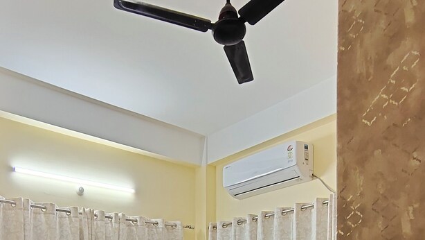 A bedroom with a bed, ceiling fan, air conditioning unit, curtains, and a small table with a plant. - Beltola Lodge - a serene homestay (Guwahati)