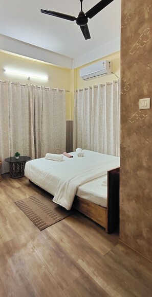 Une chambre à coucher comprenant un lit, un ventilateur de plafond, un appareil de climatisation, des rideaux et une petite table avec une plante.
