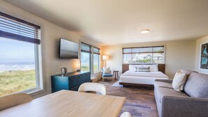 A hotel room with a large bed, a sofa, a dining table, a TV, and a view of the beach. - Manzanita Oceanfront Inn (Manzanita)