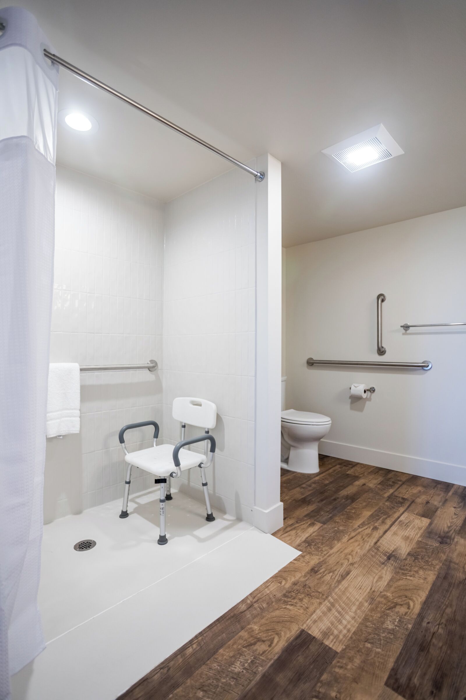 Un baño con ducha, inodoro, barra de apoyo y banco accesible para sillas de ruedas.