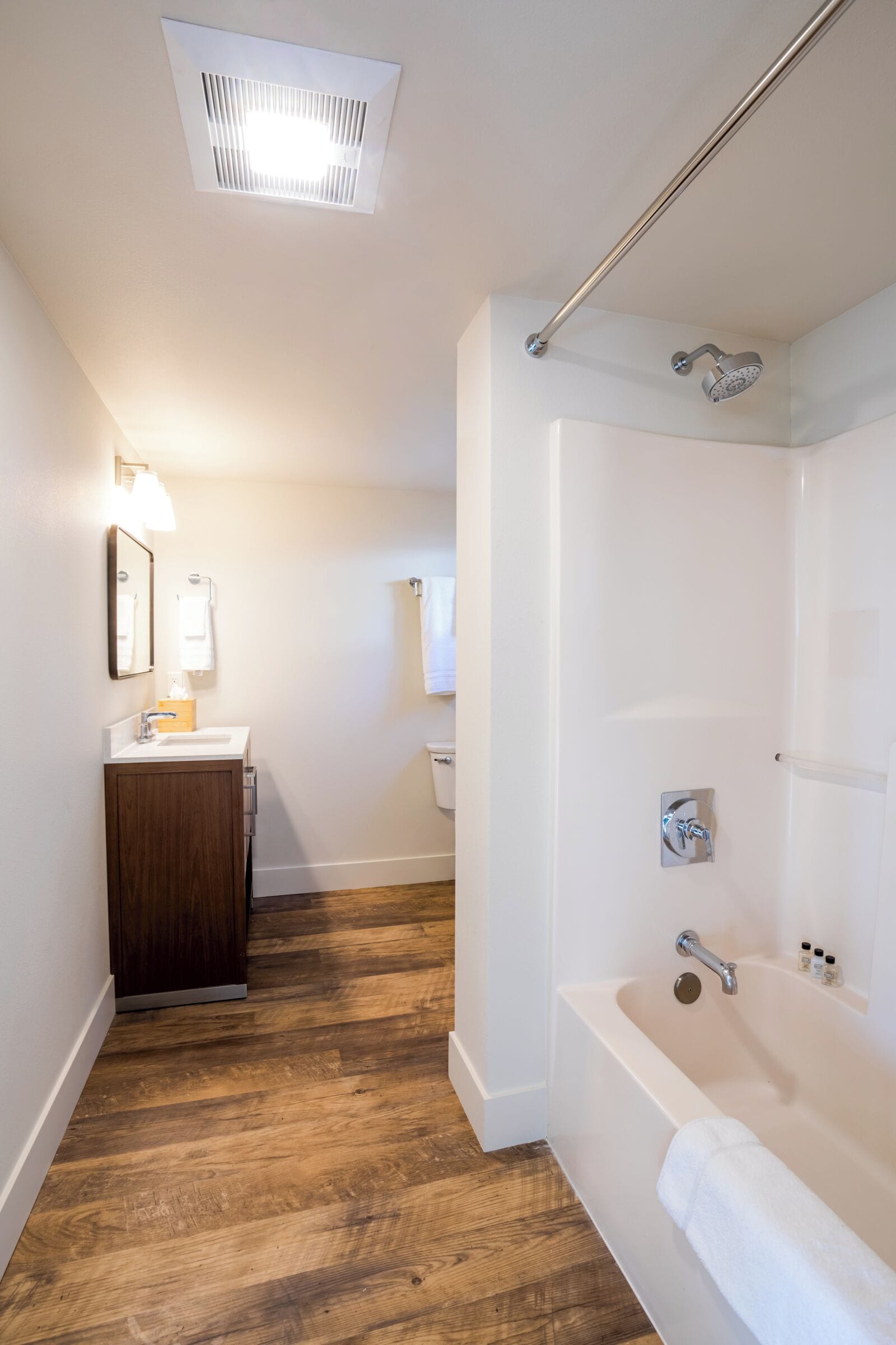 Een moderne badkamer met houten vloer, een wit ligbad, een douche met regendouche, een wastafel met spiegel en handdoek, en een plafondlamp.
