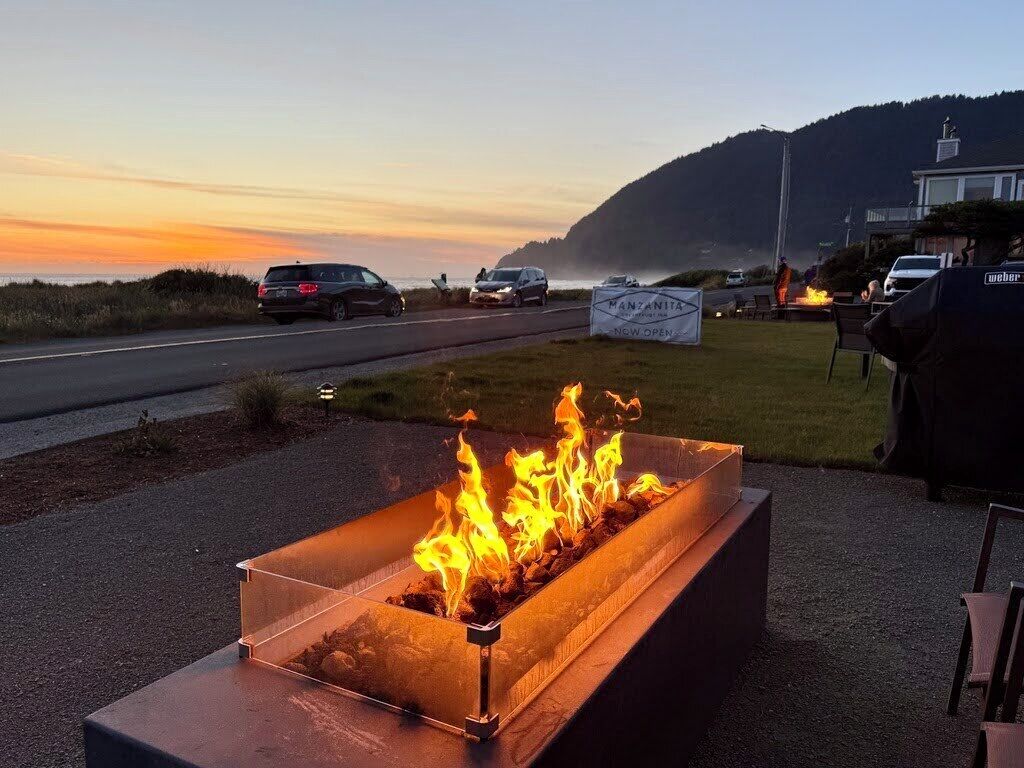 Un elemento decorativo con fuoco, un cartello che annuncia l'apertura, una strada con veicoli e una zona erbosa con sedie.