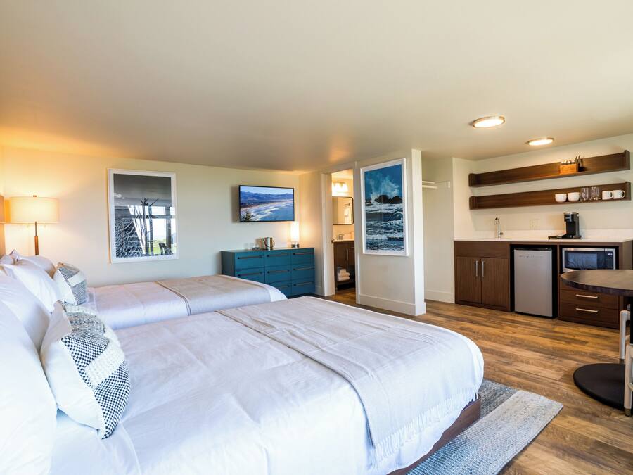 A hotel room with two beds, a desk, a chair, a kitchenette, and artwork on the walls.