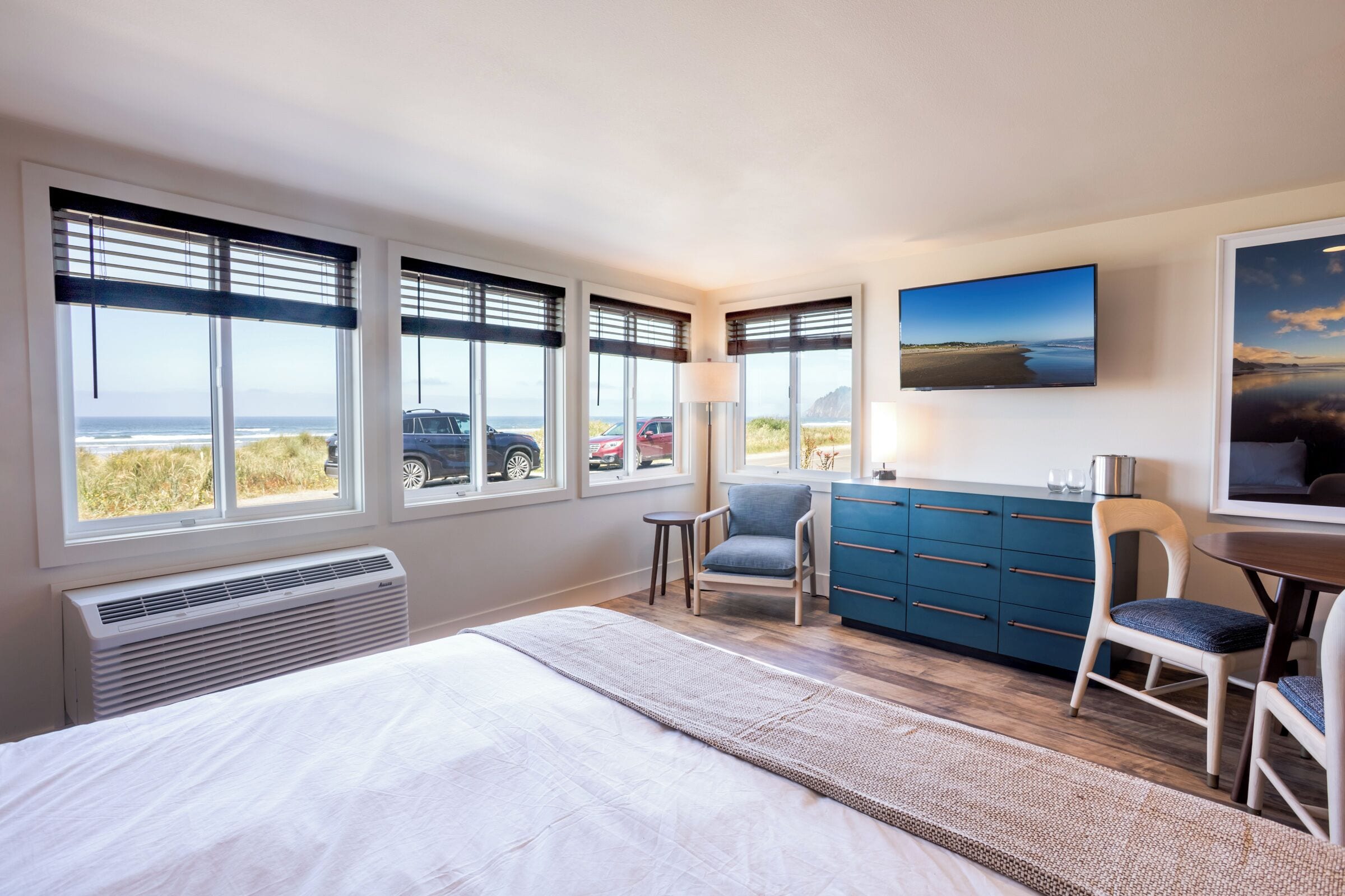 Un dormitorio con cama, escritorio, silla, aparador y ventanas amplias con vistas a la playa y coches estacionados afuera.