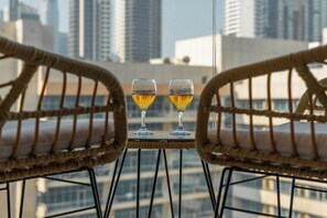 Zwei Korbstühle mit Kissen, ein kleiner Tisch und zwei Gläser eines gelblichen Getränks vor einer Stadtkulisse.