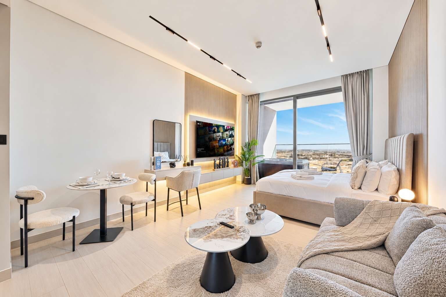A modern hotel room with a large bed, a sofa, a dining area, and a TV mounted on the wall.