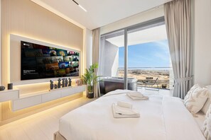 A modern hotel room with a large TV mounted on the wall displaying various channels, a large bed with neatly folded towels, a hot tub, and a balcony with a city view. - Chic Studio w/ Private Jacuzzi / Cold Plunge / Gym (Dubai)