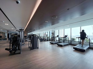 A modern gym with cardio equipment, weight machines, and large windows offering a city view. - Chic Studio w/ Private Jacuzzi / Cold Plunge / Gym (Dubai)