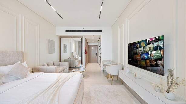 Ein modernes Hotelzimmer mit einem großen Bett, einer Sitzecke inklusive Sofa und Sesseln, einem an der Wand befestigten Fernseher und einem Flur zu weiteren Zimmern.