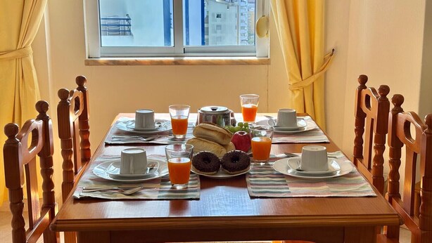 Ein Frühstückstisch ist gedeckt mit Tellern, Tassen und verschiedenen Speisen, darunter Früchte und Gebäck, in einem Raum mit Holzsesseln und Blick auf Gebäude durch ein Fenster.