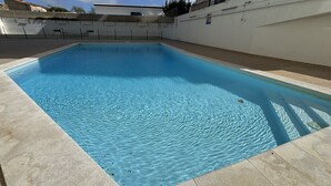 Ein rechteckiger Swimmingpool mit klarem, blauem Wasser, umgeben von einer gefliesten Liegefläche und einer weißen Wand mit einem Balkon.