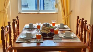 A breakfast table set with plates, cups, and a variety of food items, including fruits and pastries, in a room with wooden chairs and a view of buildings through a window.