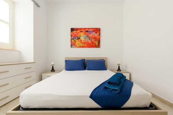 A bedroom with a bed, two bedside tables, a window, and a painting on the wall.