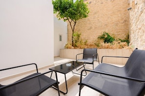 A modern outdoor seating area with a glass-top table and black chairs, surrounded by stone walls and a tree.