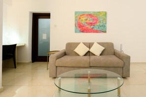 A living room with a beige sofa, two white cushions, a glass coffee table, and a colorful abstract painting on the wall.