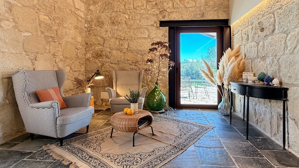Um quarto aconchegante com parede de pedra, uma poltrona xadrez, um pequeno banquinho, uma luminária, um vaso com plantas secas e vista através de uma porta.