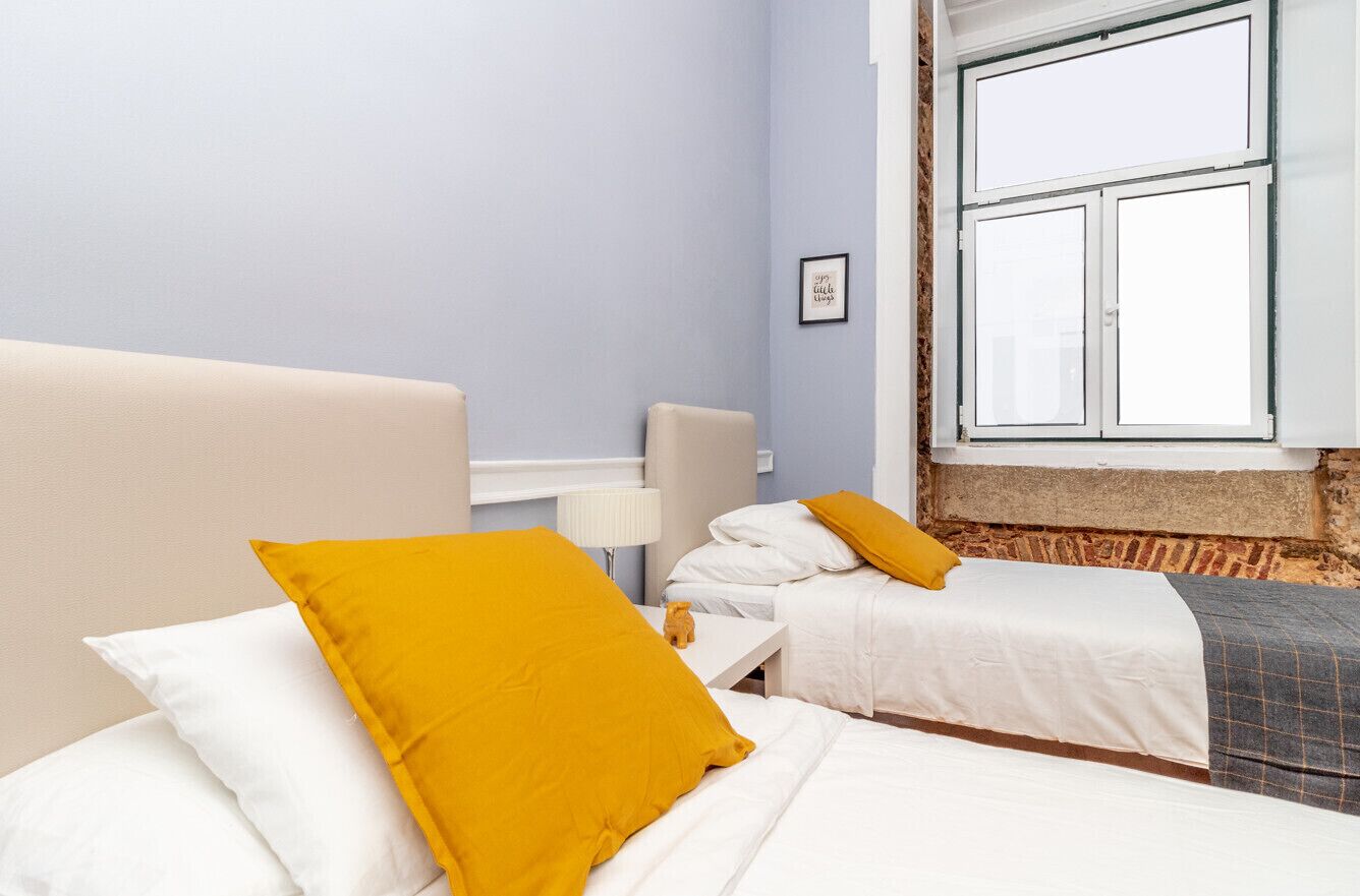 Habitación de hotel con dos camas, una con cabecera blanca y la otra con cabecera gris. Se ve una ventana con marcos blancos y vistas al exterior.