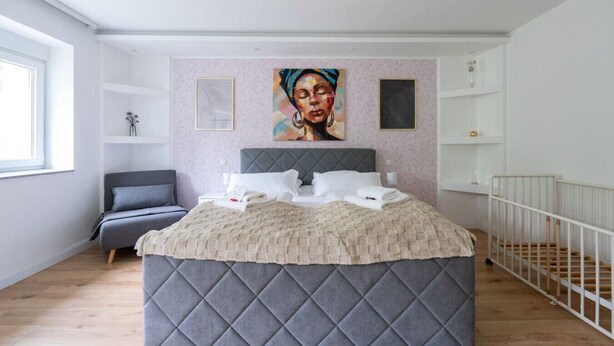 Un dormitorio moderno con una cama grande, cabecera gris, mesita de noche, escritorio pequeño, silla, ventana y barandilla blanca.