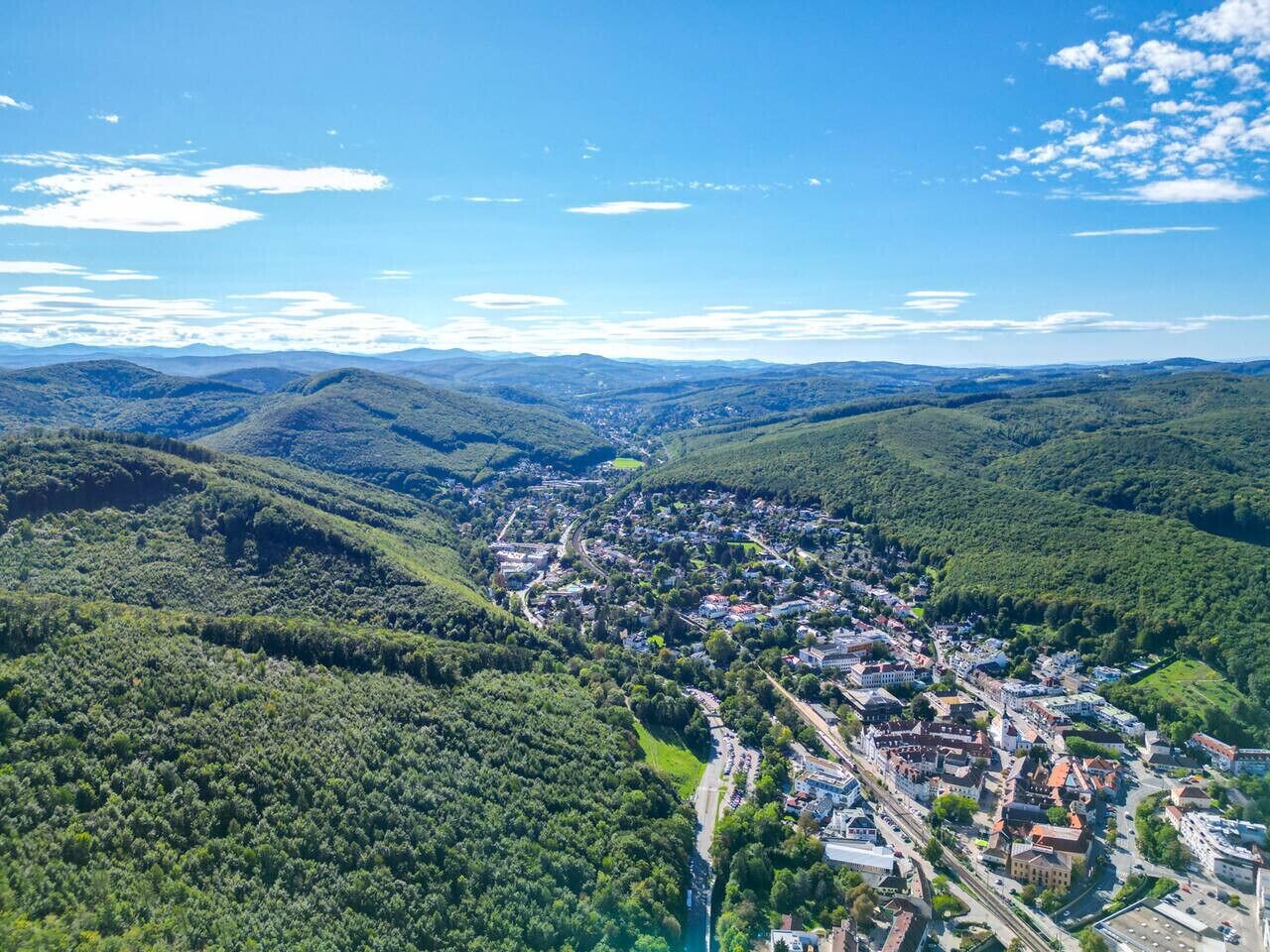 An aerial view of a town nestled in a valley with dense forested hills, a winding road, and a mix of residential and commercial buildings.