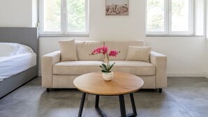 Un dormitorio moderno con cama, sofá, una mesita redonda con un jarrón de flores y dos ventanas con vistas a zonas verdes.
