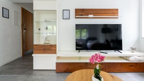 Una sala de estar moderna con una televisión de pantalla plana montada en un estante de madera, un aparador de vidrio y una mesa redonda de madera con una orquídea rosa.